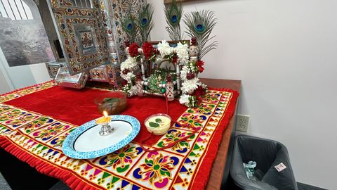 A vibrant altar with colorful decorations, flowers, and peacock feathers, featuring a lit candle and food offerings on a red cloth.