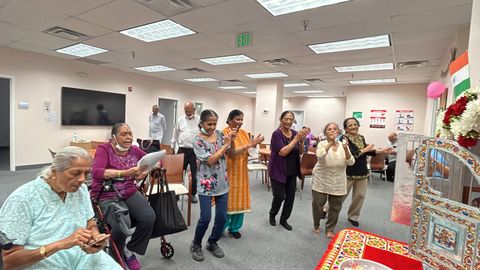 A group of people in a room, some clapping and others seated, around a puja in a community room, with the Indian flag in the background.