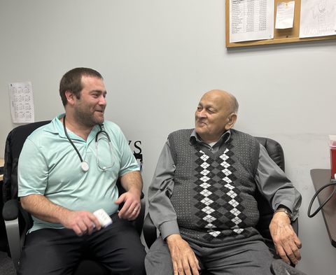 A healthcare professional with a stethoscope talks to an elderly man in an office setting, both sitting and smiling.