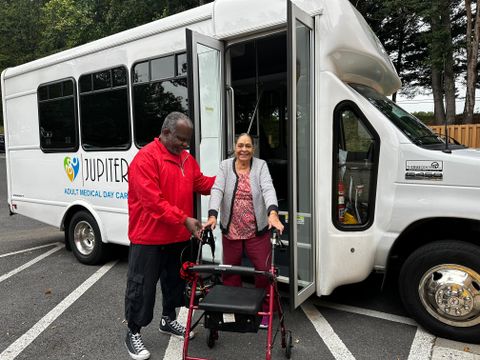 A man assists a woman with a walker as she exits a Jupiter Adult Medical Day Center van parked in a lot.