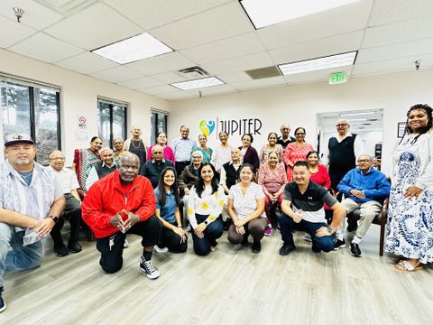 A group photo of Jupiter Care staff and participants. People are posed in a semicircle in the main room of the day center, with the Jupiter Care logo visible on the back wall behind them. The front row is kneeling and the back row is standing. The Center’s founder, Shivangi Patel, kneels in the center of the front row.