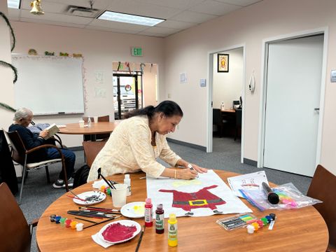 A woman paints a festive design on paper at a table with art supplies, while another person reads in the background of a community room.
