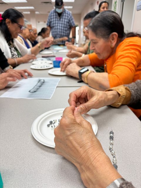 A closeup of a participant's hands as they add beads to a cord. Other participants are visible in the background, also stringing beads.