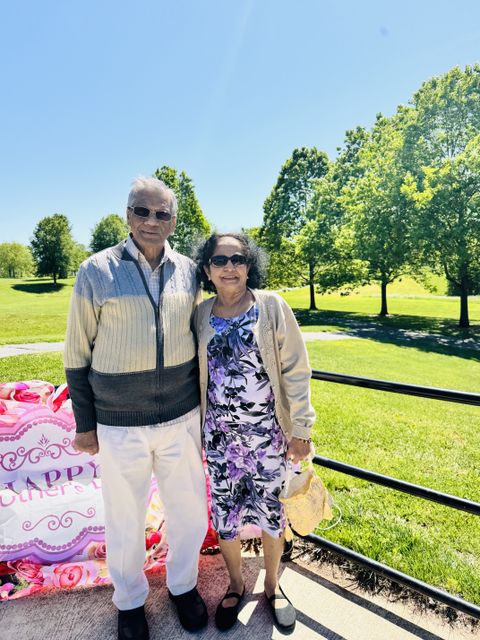 Elderly couple smiling outdoors on a sunny day, standing on a pathway with green trees and a bright sky in the background.