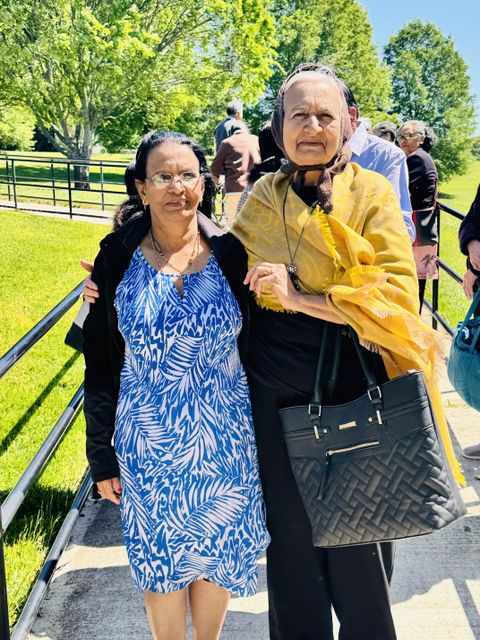 Two women standing together outdoors, one in a blue patterned dress, the other in a yellow shawl, with trees and people in the background.