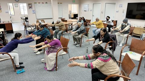 A teacher leads a stretching activity with a group of participants in a community room. Everyone is leaning forward in their chairs with their arms outstretched.
