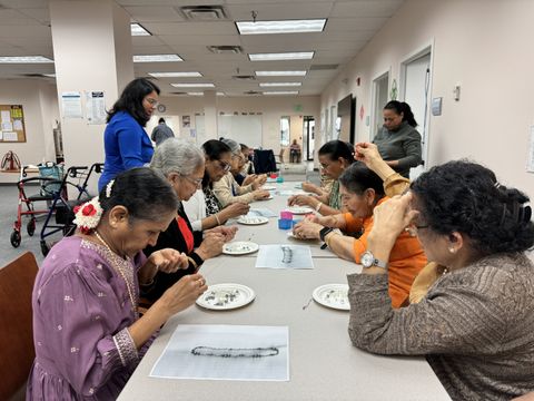 A group of participants create beaded crafts at a long table in a community room.