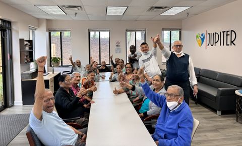 A group of elderly people and caregivers smiling and raising their hands in a bright room at the Jupiter Care Medical Day Center.