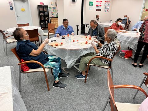Four participants sit around a table painting wooden crafts in a community room.