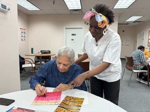 An older woman draws on pink paper at a table while another woman stands beside her, offering guidance in a well-lit room.