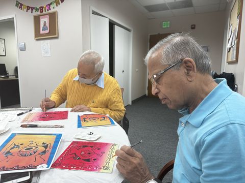 Two elderly men focused on painting colorful, abstract designs at a table in a room with birthday decorations.