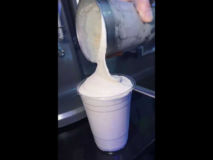 A Burgers On B creamy milkshake being poured from a metal container into a clear plastic cup, with a dark background.
