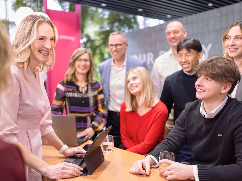 Gruppe von Mitarbeitenden bei einer gemeinsamen Veranstaltung, eine Mitarbeiterin steht vor einem Schreibtisch mit einem Tablet, weitere Mitarbeitende sitzen und stehen vor ihr und hören ihr zu.