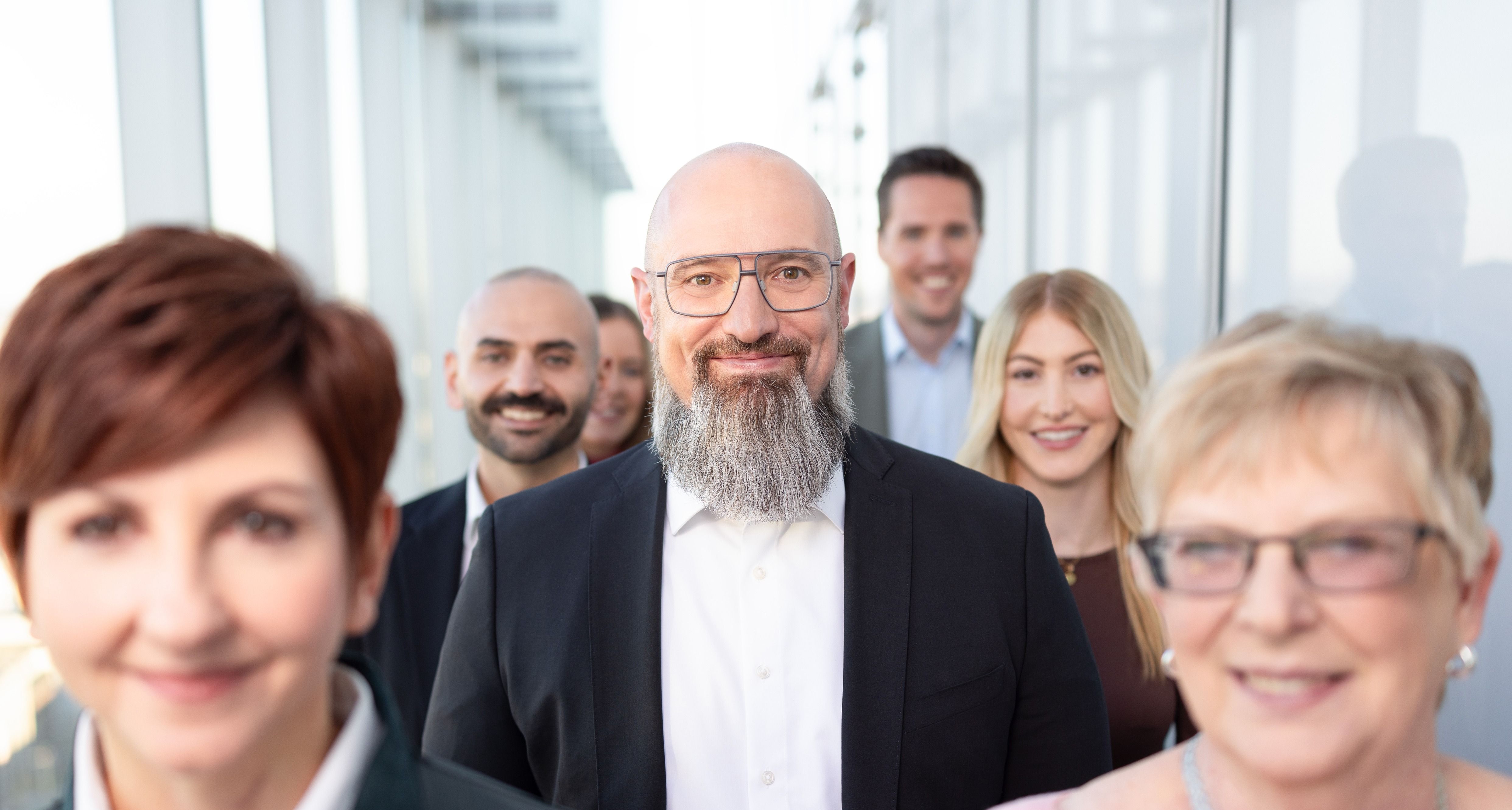 Eine Gruppe verschiedener Mitarbeitenden steht gemeinsam und hintereinander in einer Gruppe in einem modernen Arbeitsumfeld.