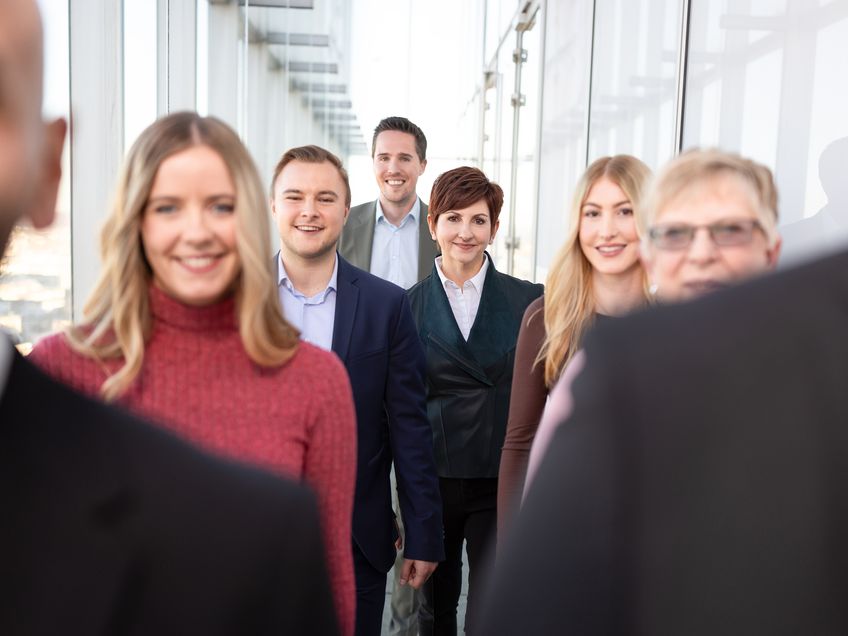 Verschiedene Mitarbeitende stehend hintereinander zusammen in einem gläsernen Gang.