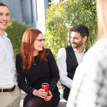 Drei Mitarbeitende stehen und sitzen zusammen, lachen und tauschen sich aus.
