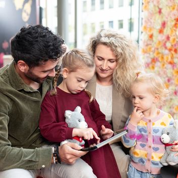 Zwei Erwachsene lesen gemeinsam mit zwei Kindern ein Buch, sitzend auf einer Couch.