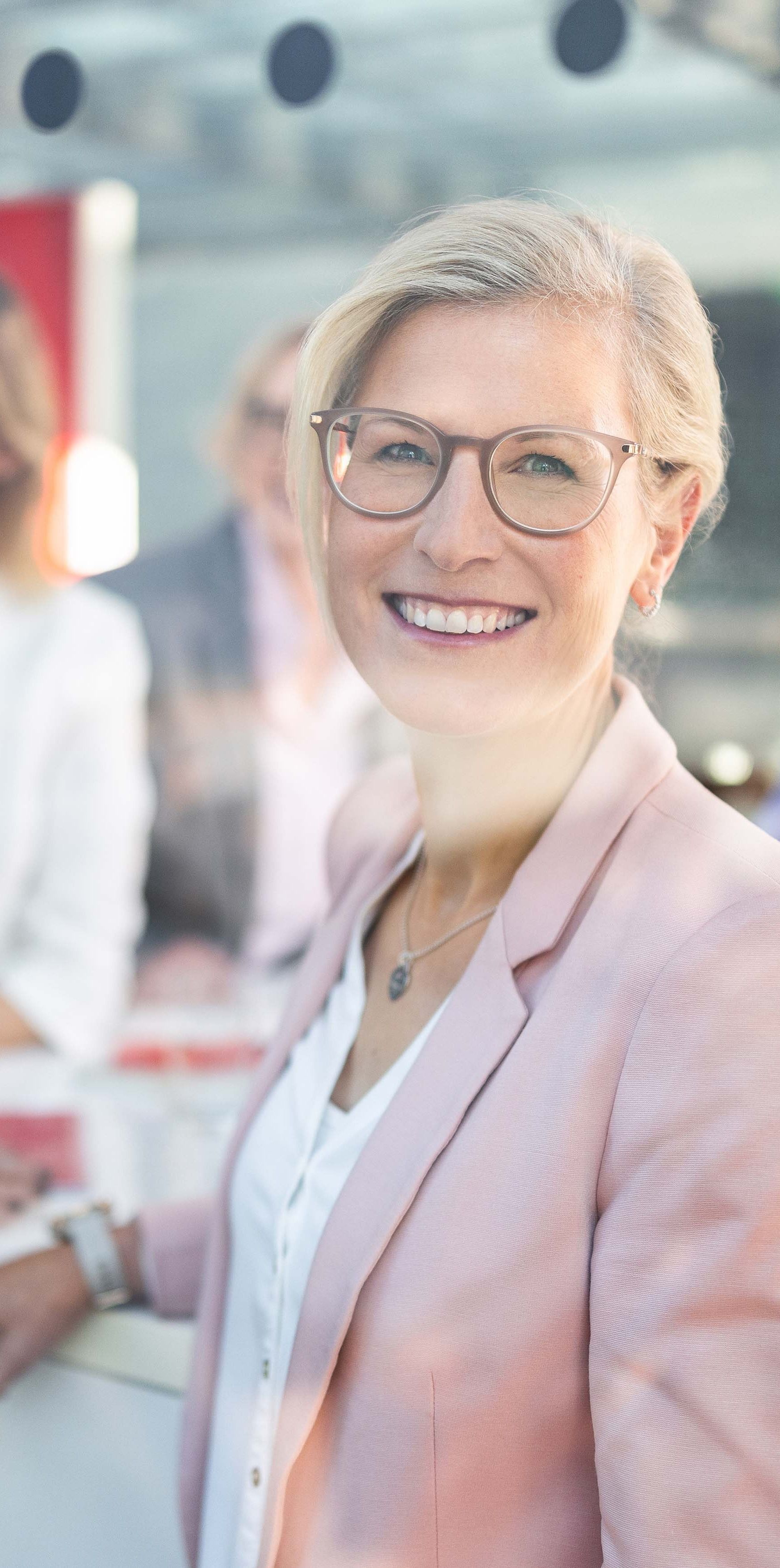 Mitarbeiterin mit Brille lächelt in einem modernen Arbeitsumfeld mit vier dahinterstehenden Kolleginnen in die Kamera
