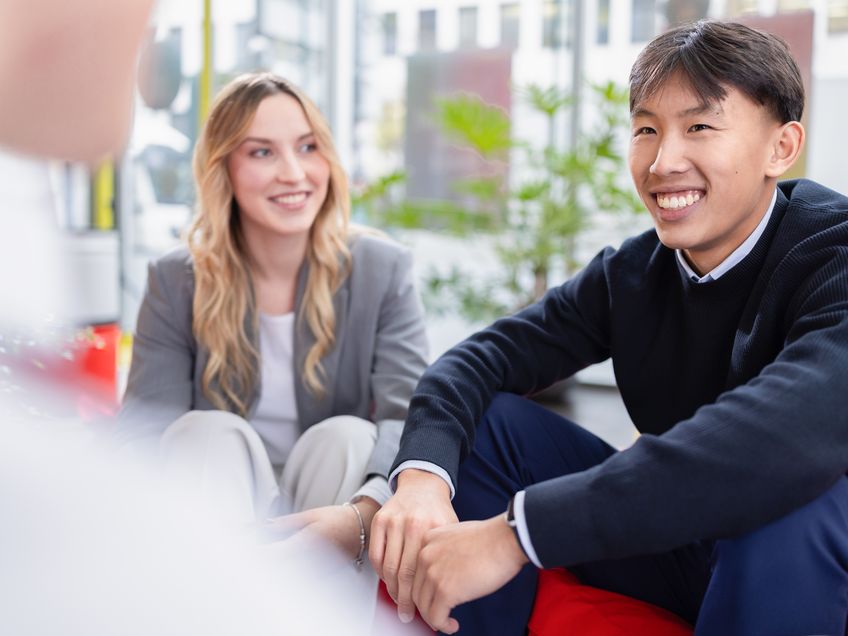 Drei Mitarbeitende sitzen gemeinsam in einem modernen Arbeitsumfeld und tauschen sich aus. Eine Person sitzt mit dem Rücken zur Kamera und ist nicht erkennbar.
