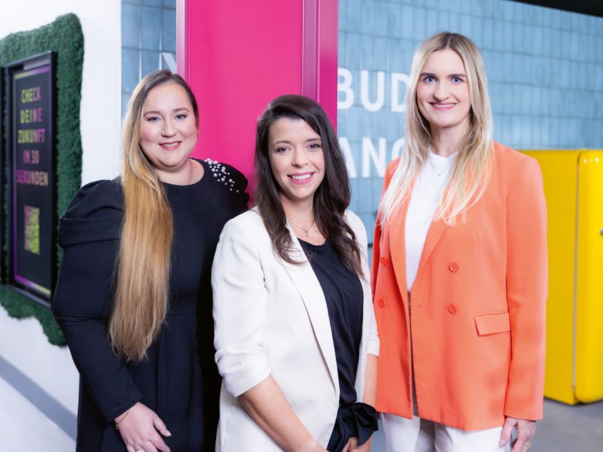 Drei Mitarbeiterinnen, die Female Finance gegründet haben, stehen zusammen in einem bunten Büroteil.