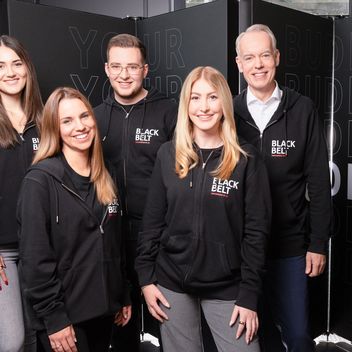 Mitarbeitende stehen alle gemeinsam zusammen, tragen einen schwarzen Hoodie mit dem Logo unsere Black Belt Programms.