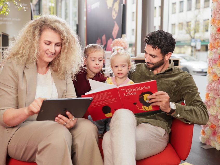 Mitarbeiterin sitzt mit ihren Kinder und ihrem Man gemeinsam auf einer Couch. Der Mann liest den Kindern aus einem Kunderbuch vor und die Frau sitzt direkt daneben und zeigt etwas auf ihrem Tablet.