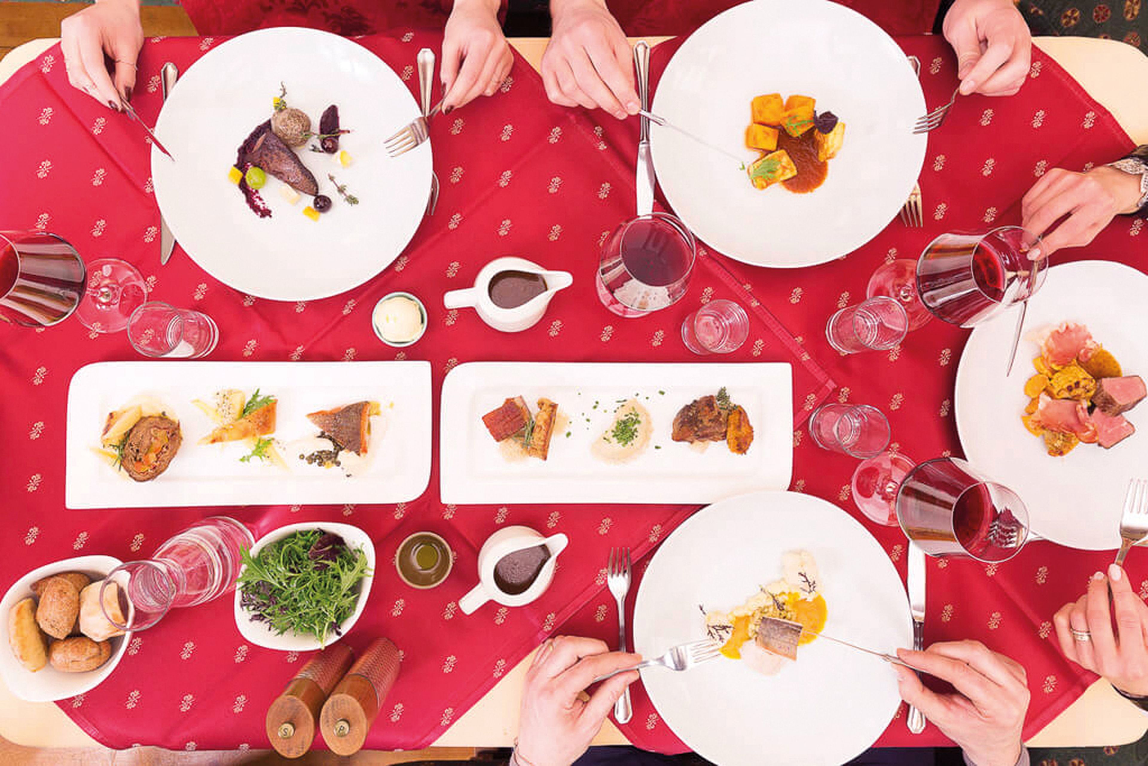 Luftaufnahme eines Esstisches im Restaurant Schloss Lerchenhof mit Menschen, die ein Gourmet-Essen genießen. Auf einer roten Tischdecke stehen elegant angerichtete Gerichte und Gläser mit Rotwein.