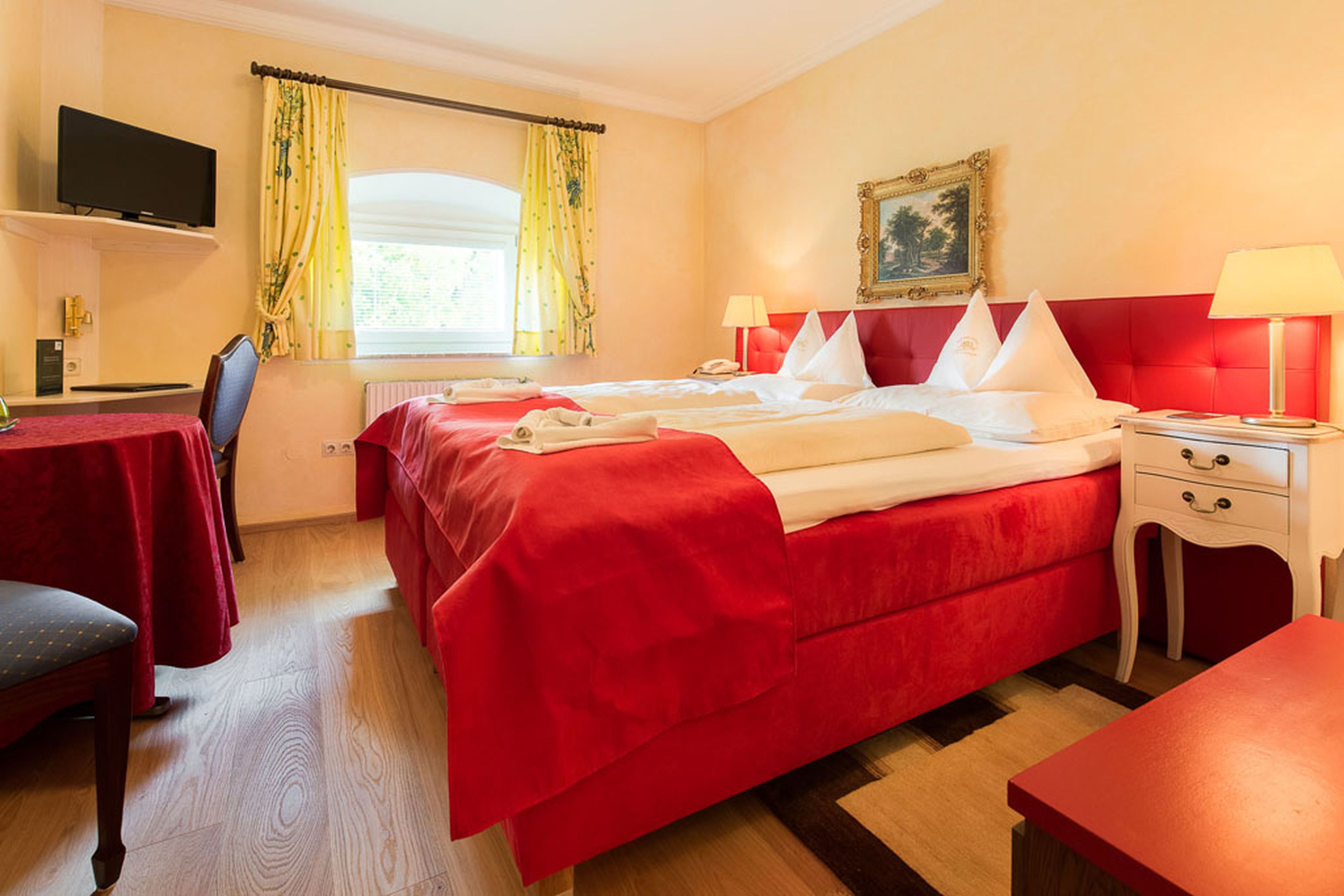 Gemütliches Hotelzimmer mit Doppelbett, roter Tagesdecke, gelben Vorhängen, Wandkunst, Fernseher und warmer Beleuchtung durch Nachttischlampen.