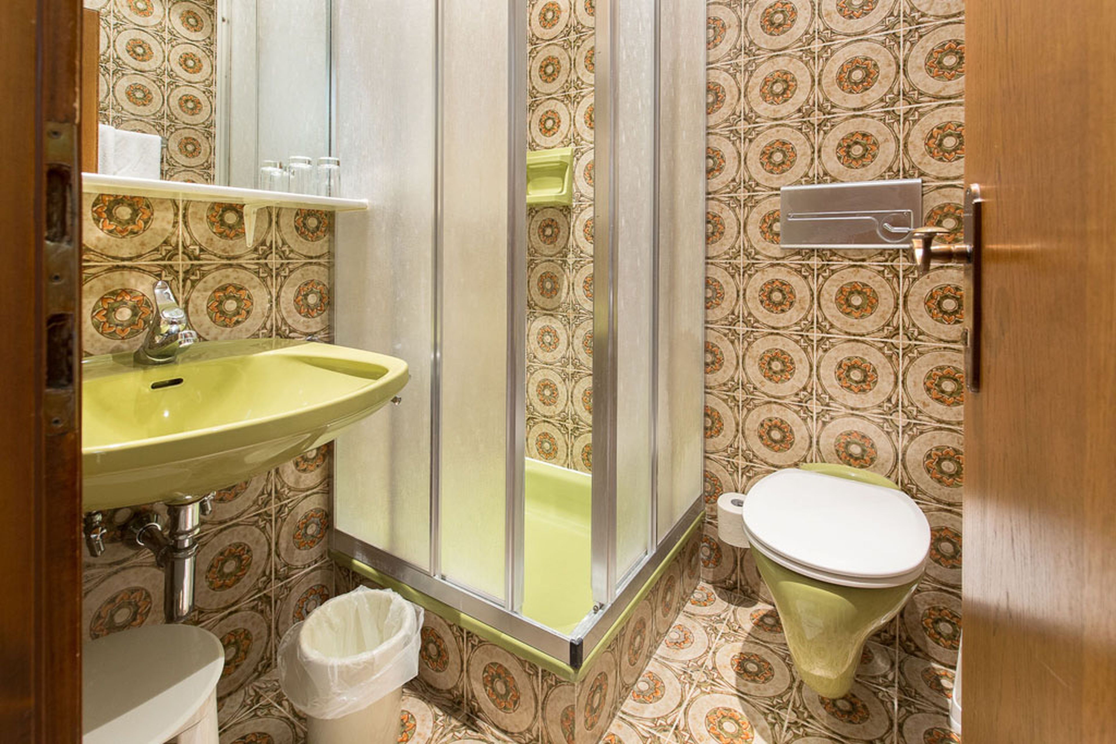 Retro-Badezimmer mit gemusterten Fliesen, grünem Waschbecken und Toilette, geschlossener Dusche und einem kleinen Regal mit Gläsern und einem Handtuch.