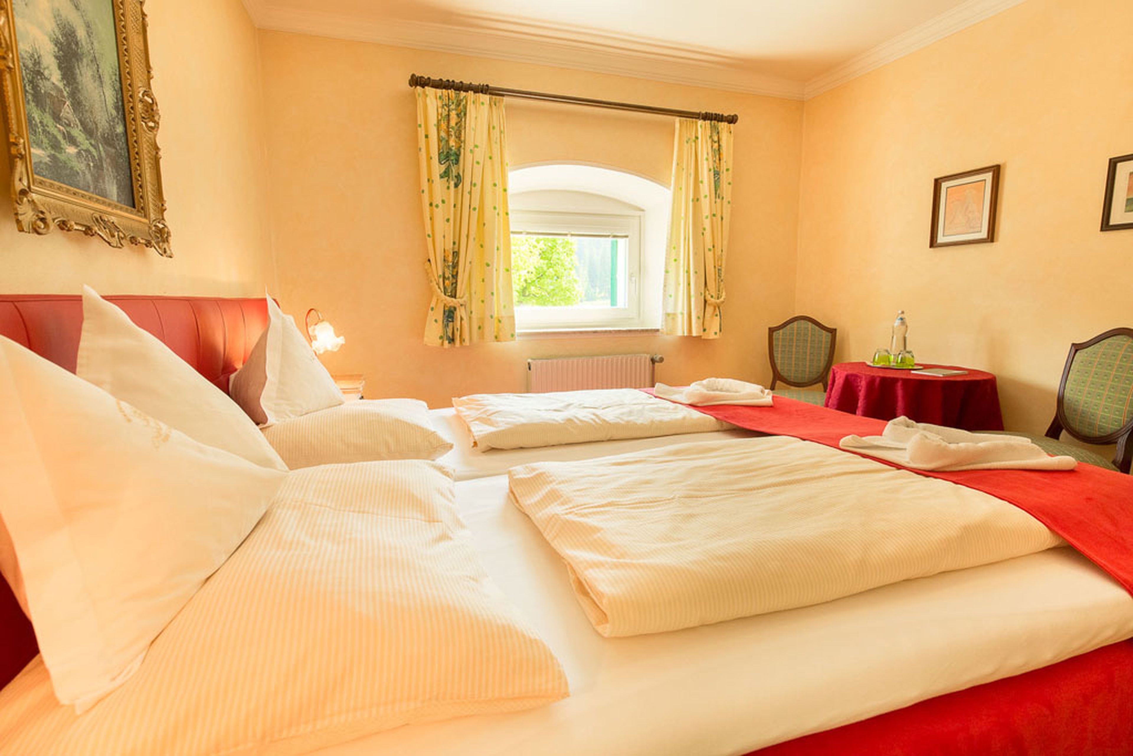 Cozy bedroom with a double bed, red accents, a window with yellow curtains, two chairs, a small table, and framed art on the walls.