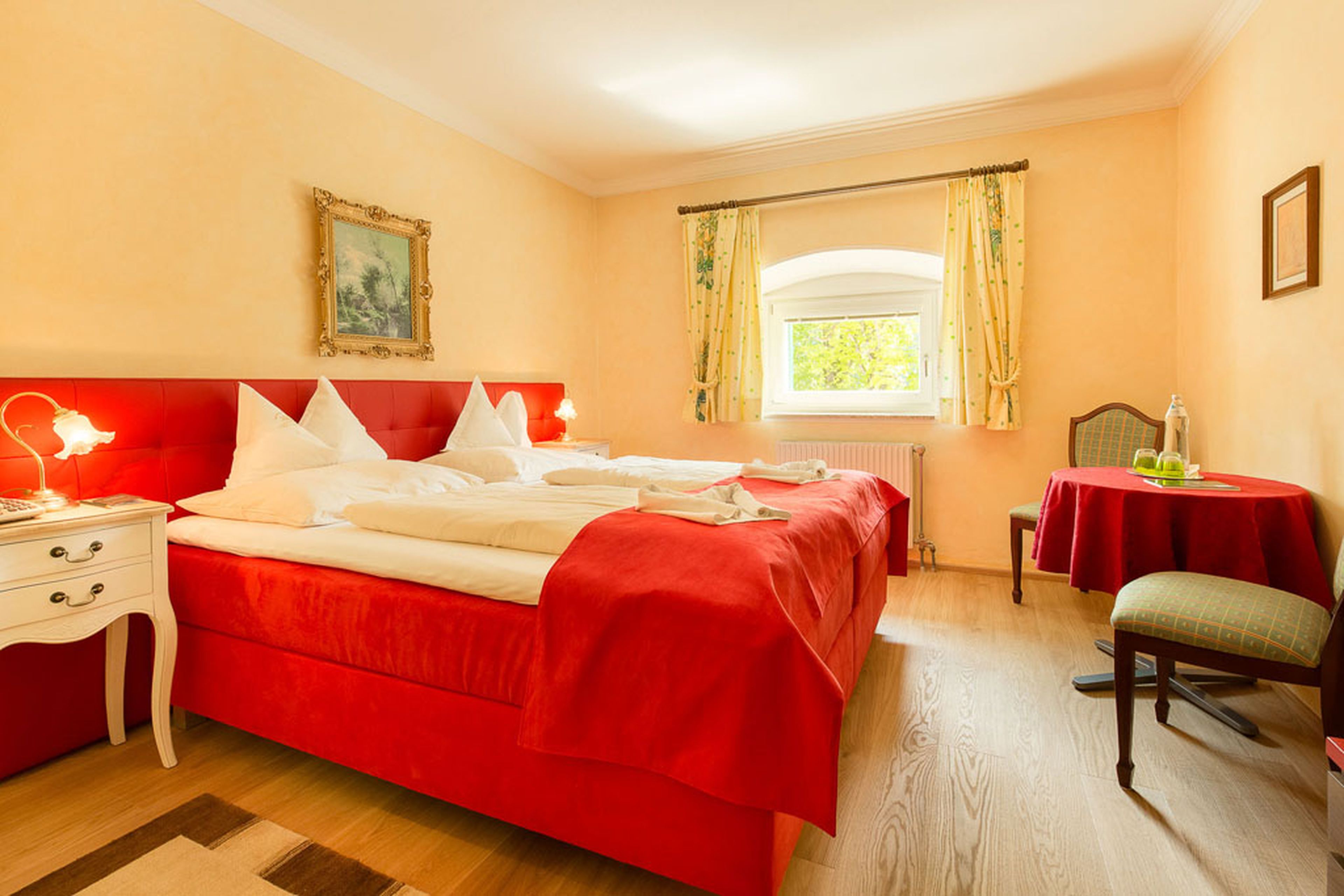 Gemütliches Schlafzimmer mit einem Doppelbett, roter Bettwäsche, Nachttischen mit Lampen, einem runden Tisch mit Stühlen und einem Fenster mit gelben Vorhängen.