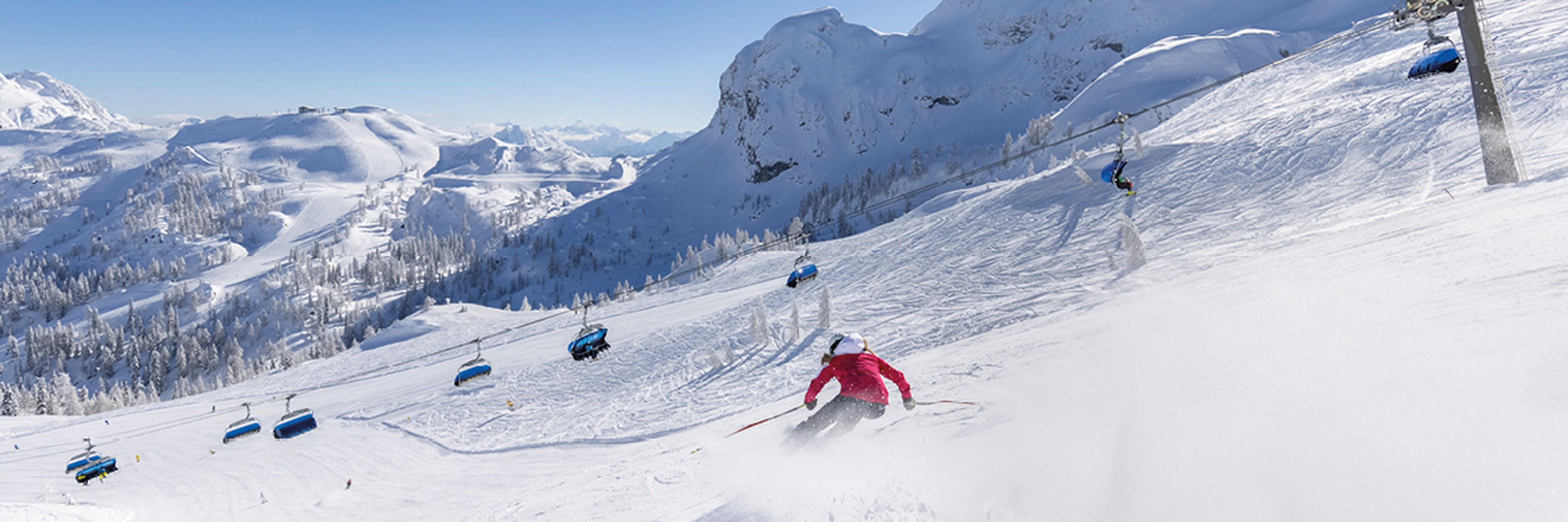 Ein Skifahrer in einer roten Jacke fährt einen verschneiten Berghang hinunter, im Hintergrund sind Sessellifte und schneebedeckte Gipfel unter einem strahlend blauen Himmel zu sehen.