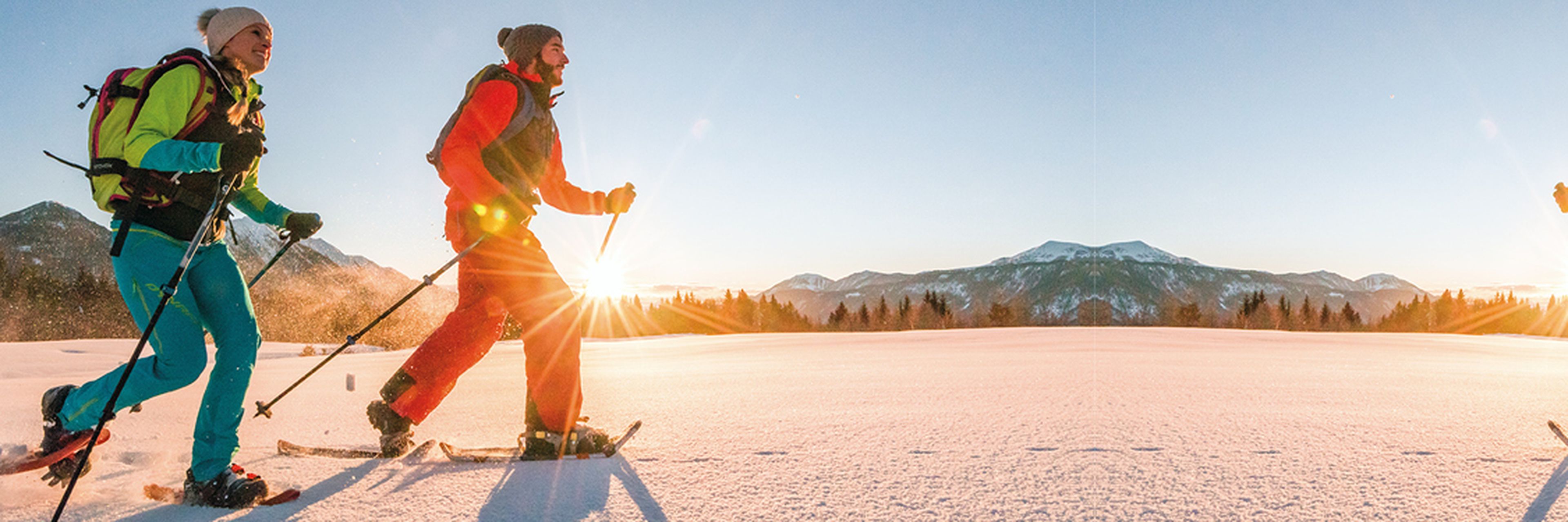 Zwei Menschen beim Schneeschuhwandern an einem sonnigen Tag, mit Bergen im Hintergrund und der tief stehenden Sonne am Horizont, die einen warmen Schein verbreitet.