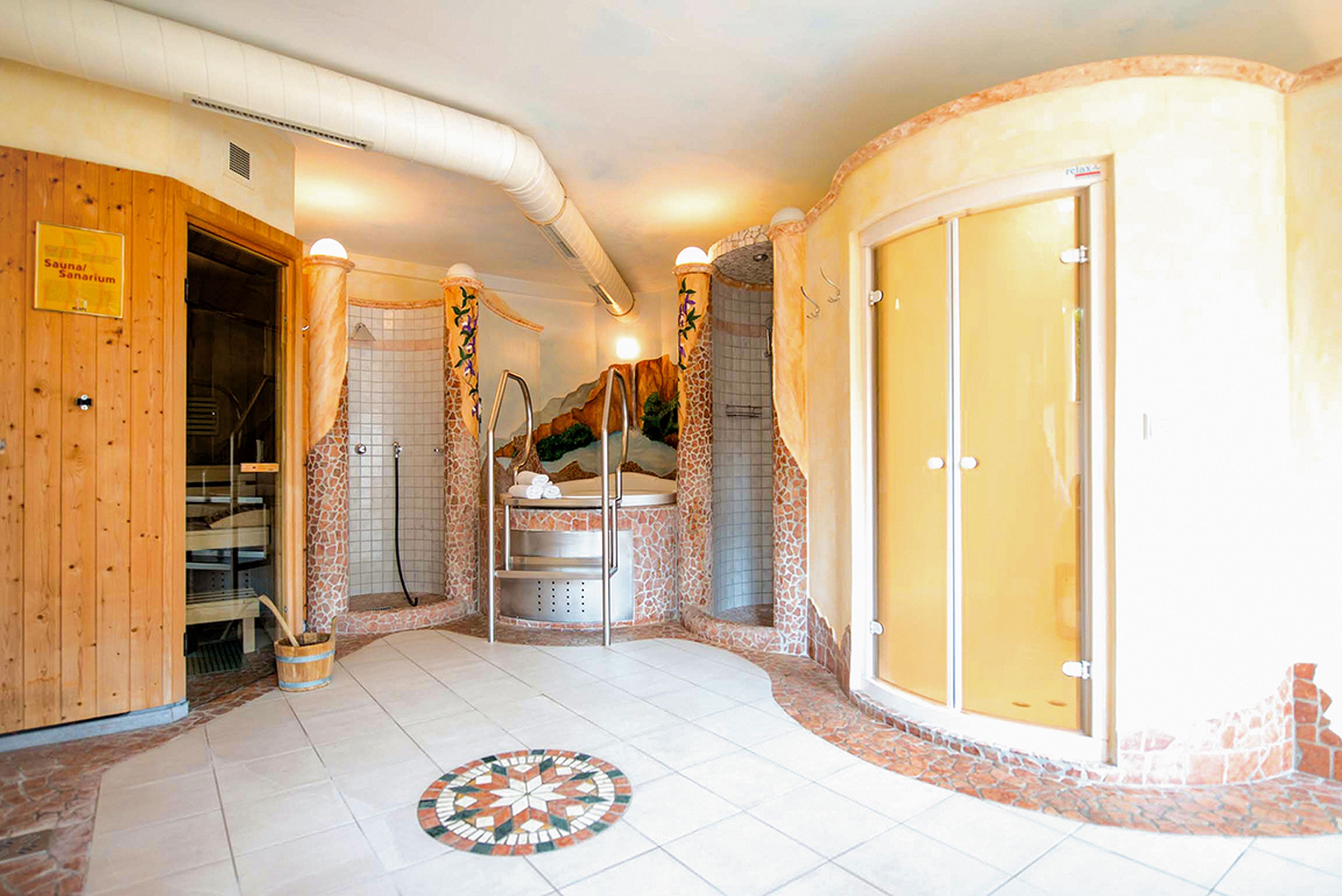 Ein Spa-Raum mit einer Sauna, zwei Duschen und einem runden Whirlpool. Die Einrichtung ist in warmen Tönen gehalten und mit Mosaikfliesen auf dem Boden und an den Wänden verziert.