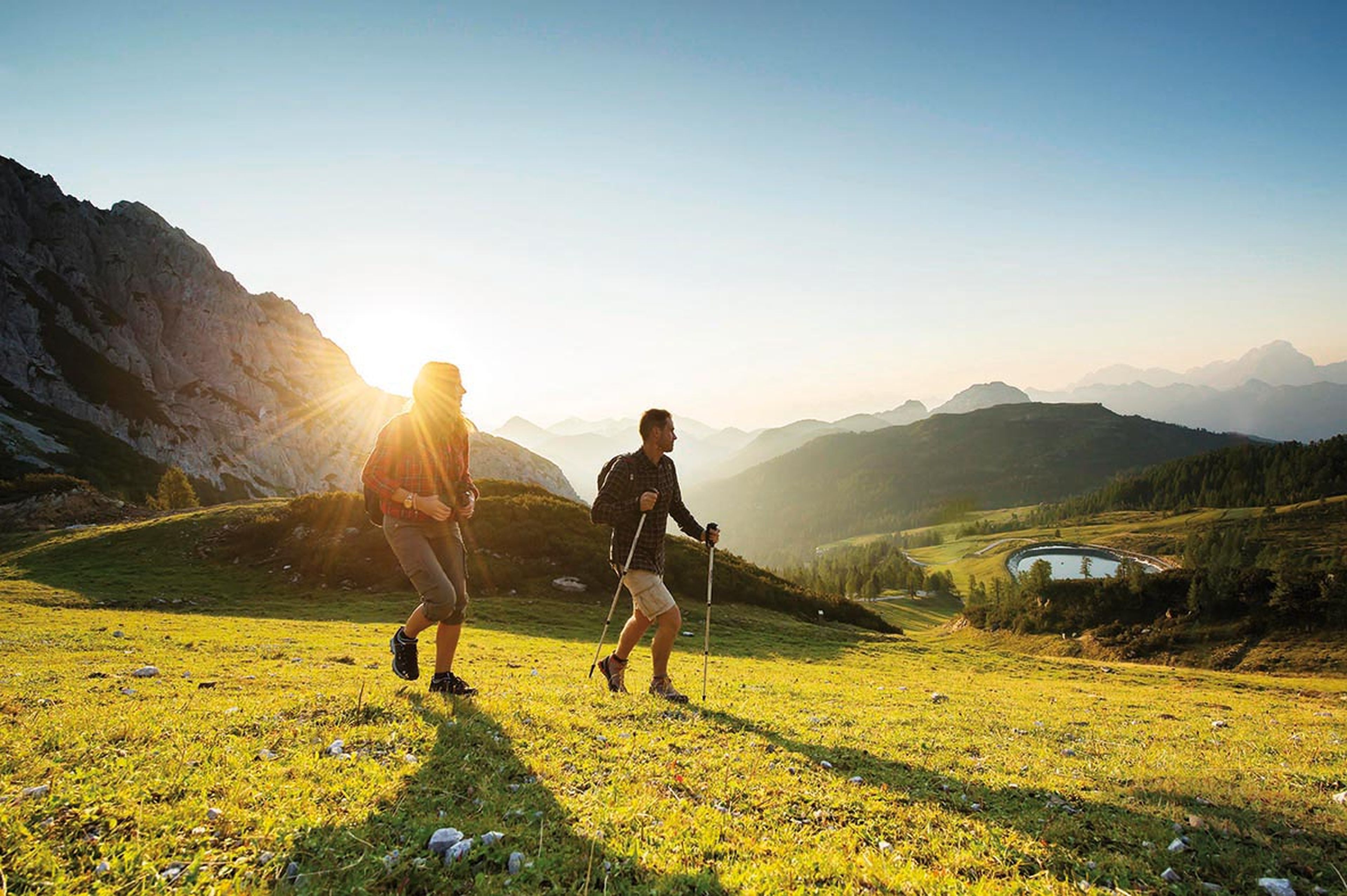 Zwei Personen wandern bei Sonnenaufgang auf einem grasbewachsenen Hügel, im Hintergrund sind Berge und ein kleiner See zu sehen.