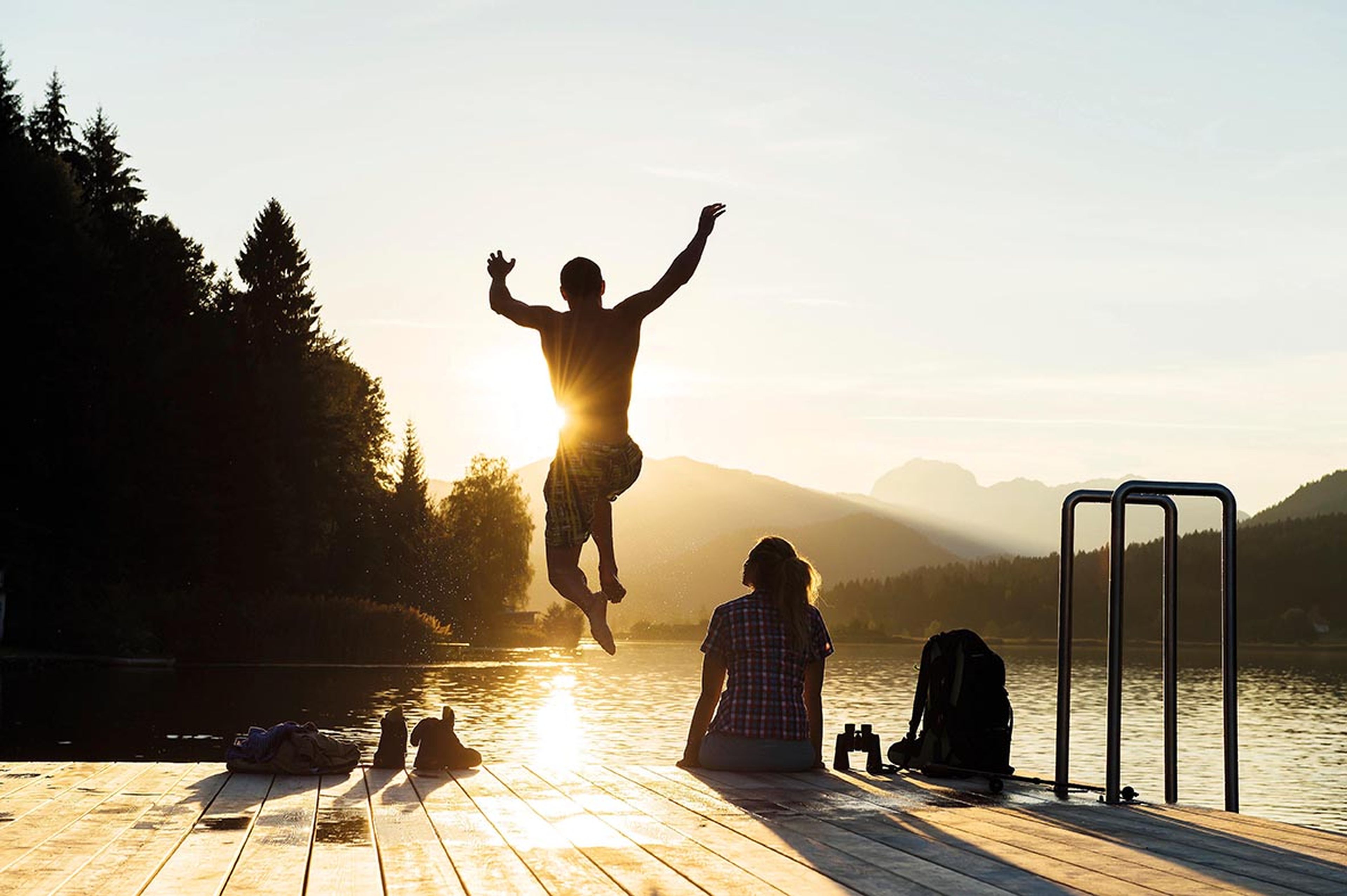 Eine Silhouette springt bei Sonnenuntergang von einem Steg am Seeufer, während eine andere Person zusieht. Im Hintergrund sind bewaldete Hügel und Berge zu sehen.