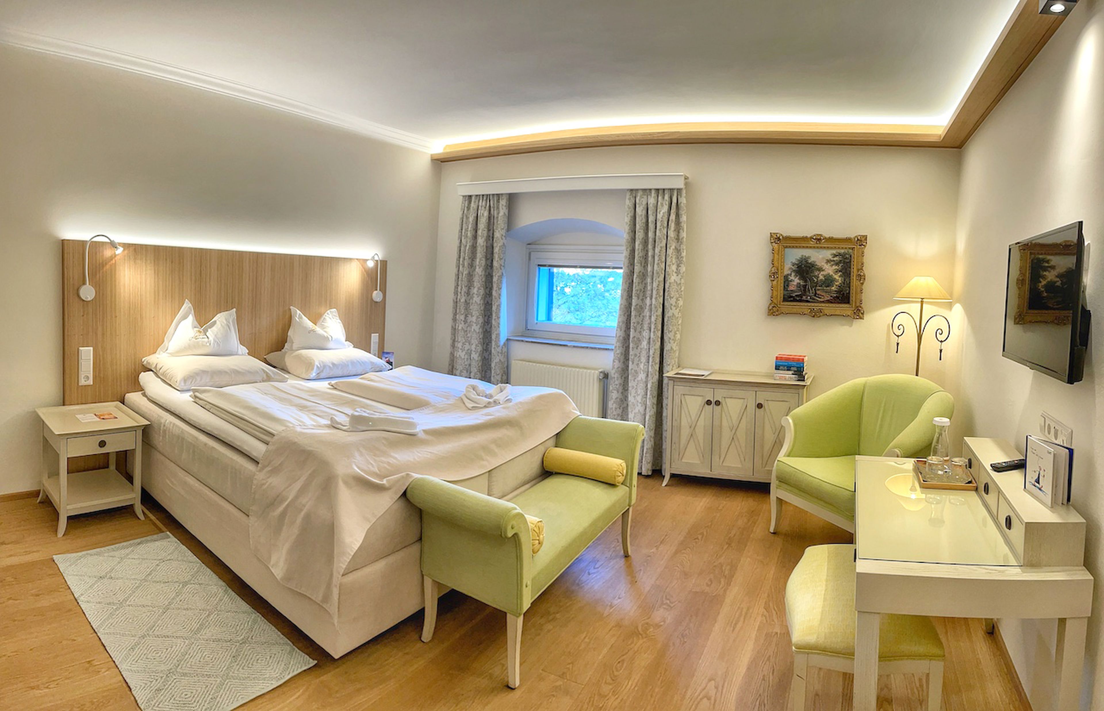 Gemütliches Hotelzimmer mit Doppelbett, grünen Stühlen, kleinem Schreibtisch, Wandkunst und sanfter Beleuchtung. Ein Fenster mit Vorhängen bietet einen ruhigen Ausblick.