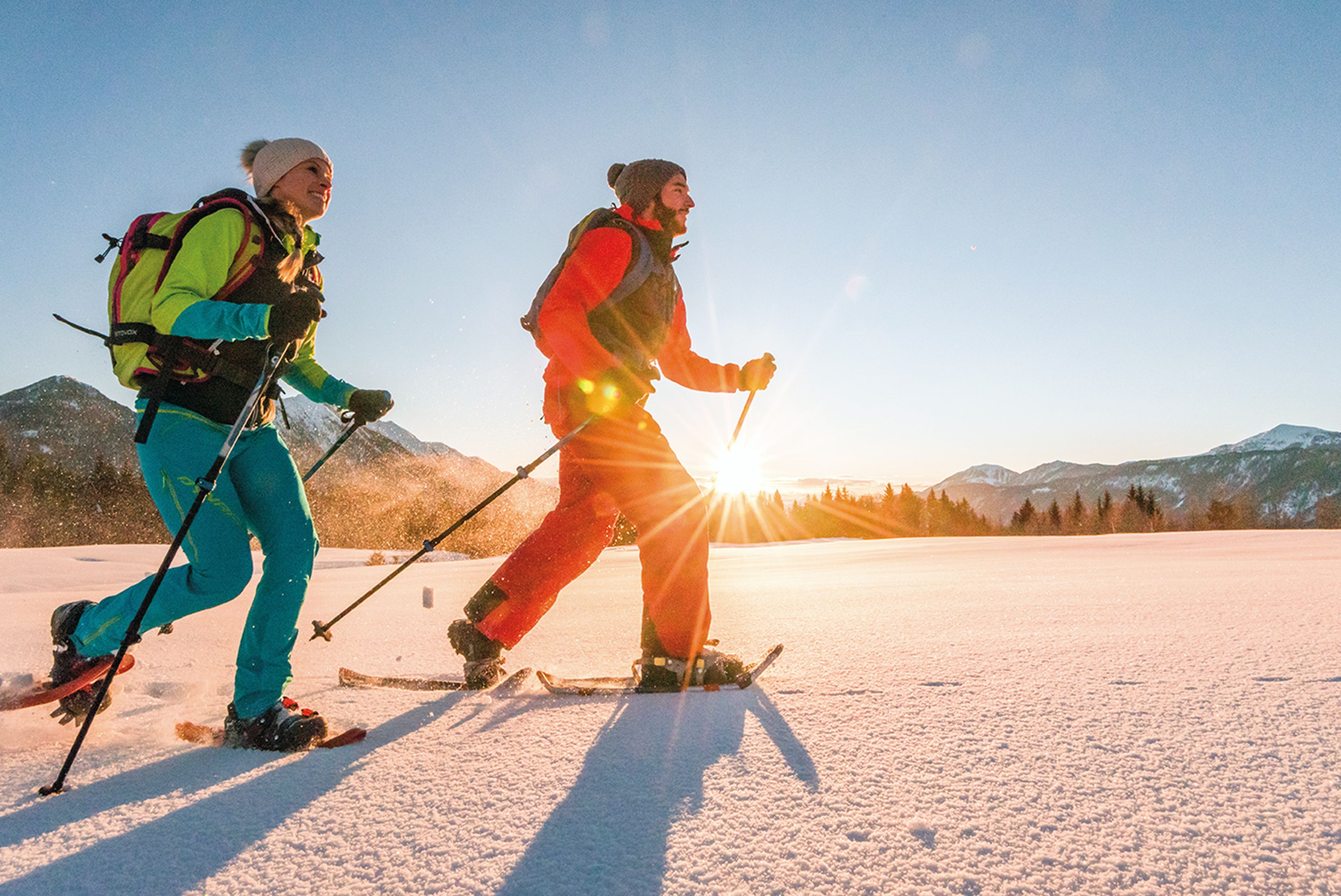 Zwei fröhliche Schneeschuhwanderer, im Hintergrund der Sonnenaufgang hinter den Bergen und Wäldern