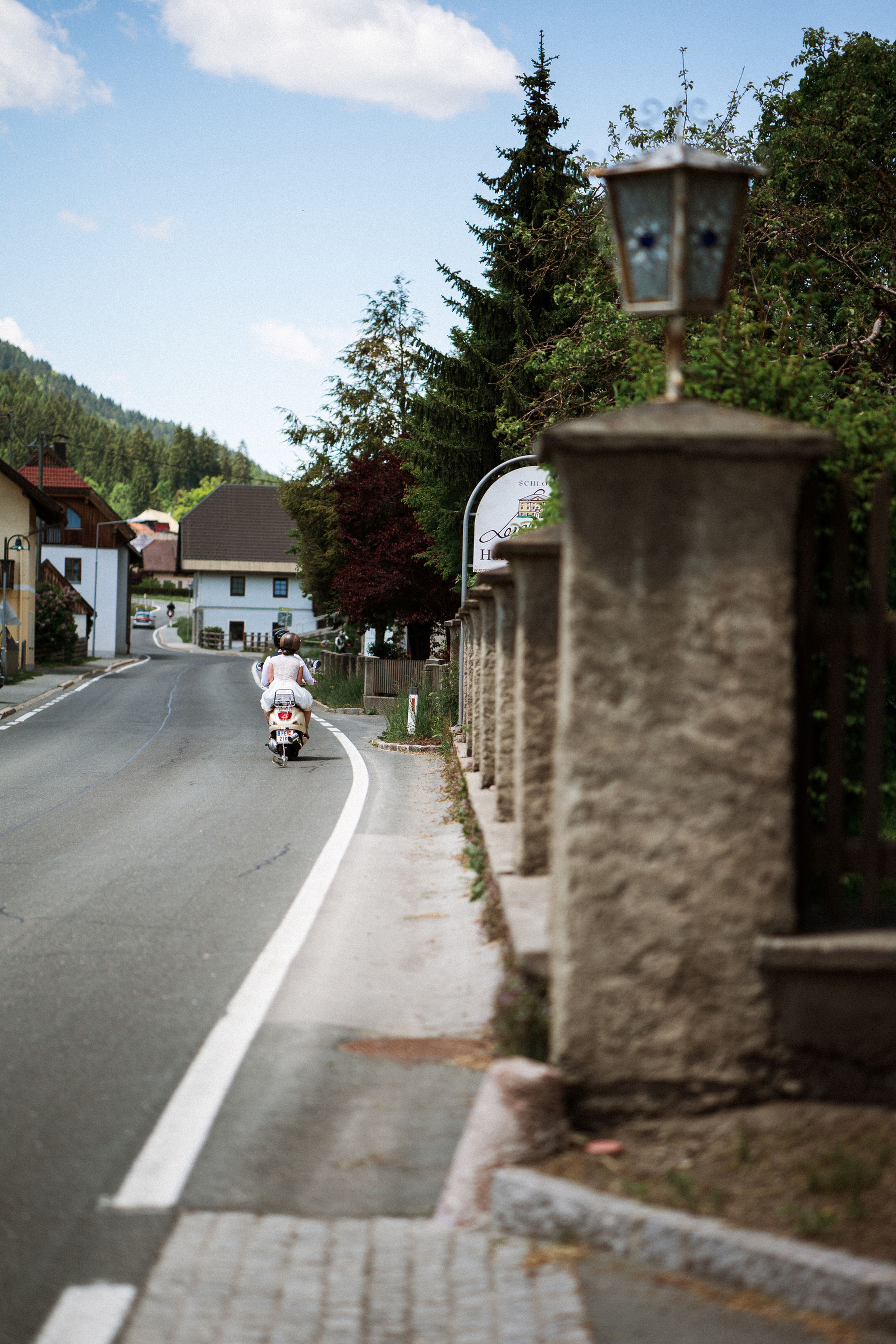 Person riding a scooter on a quiet, winding road in a rural area with houses, trees, and a stone wall under a partly cloudy sky.