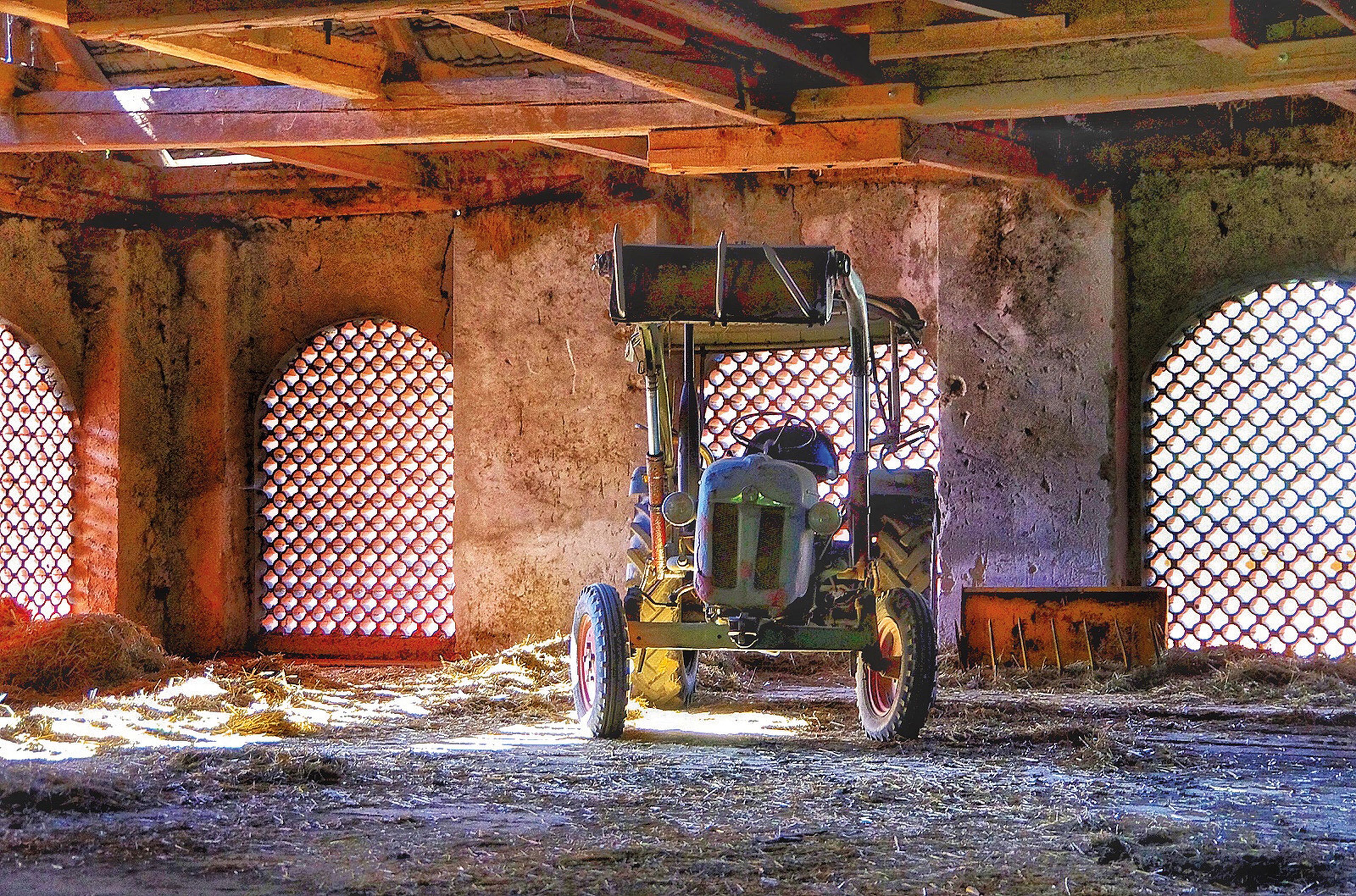 Ein Oldtimer-Traktor parkt in einer rustikalen Scheune mit gewölbten, gitterförmigen Fenstern, die sanftes Licht auf den strohbedeckten Boden werfen.
