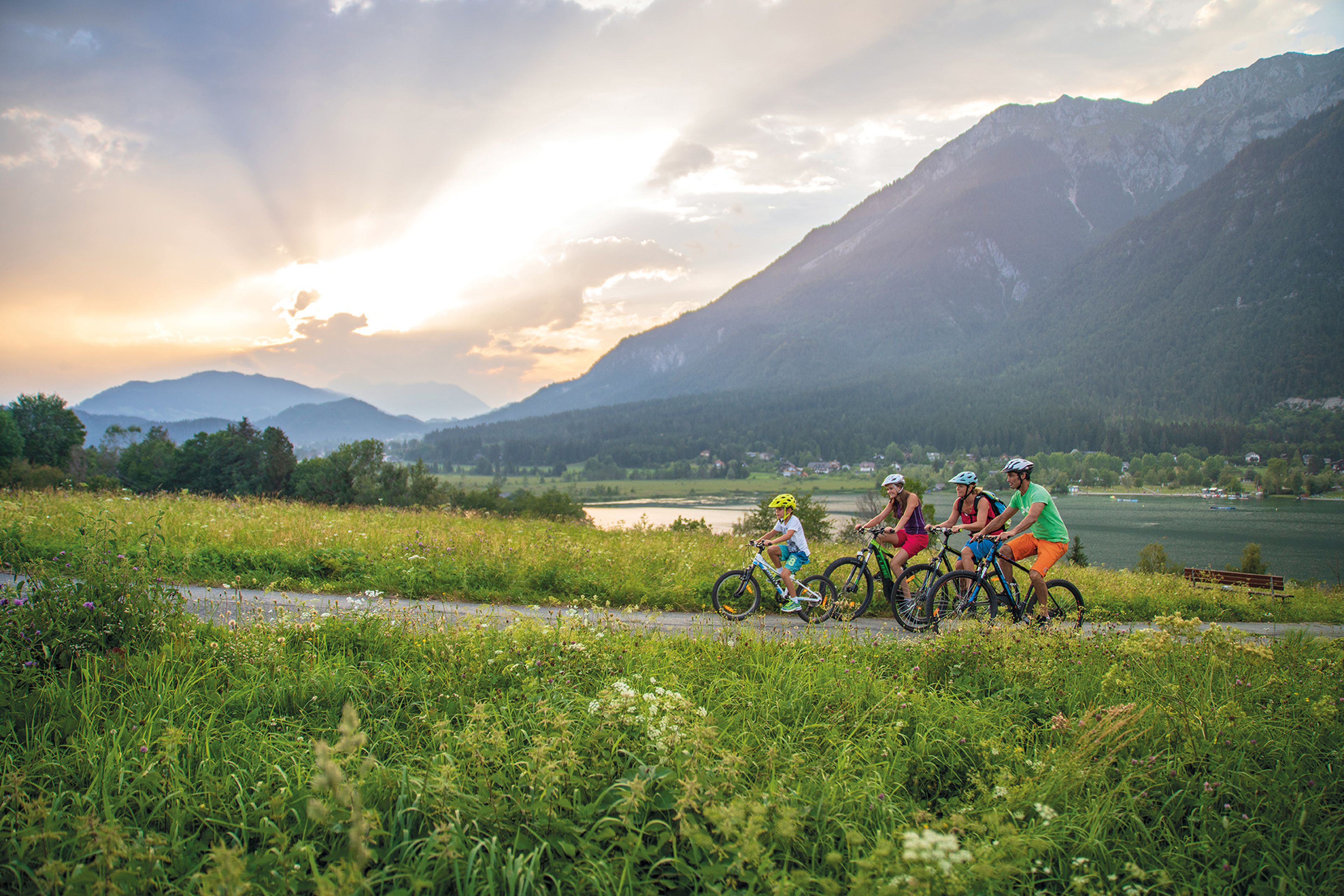 Eine vierköpfige Familie radelt auf einem Weg durch eine üppige grüne Wiese, mit Bergen und einem dramatischen Sonnenuntergangshimmel im Hintergrund.