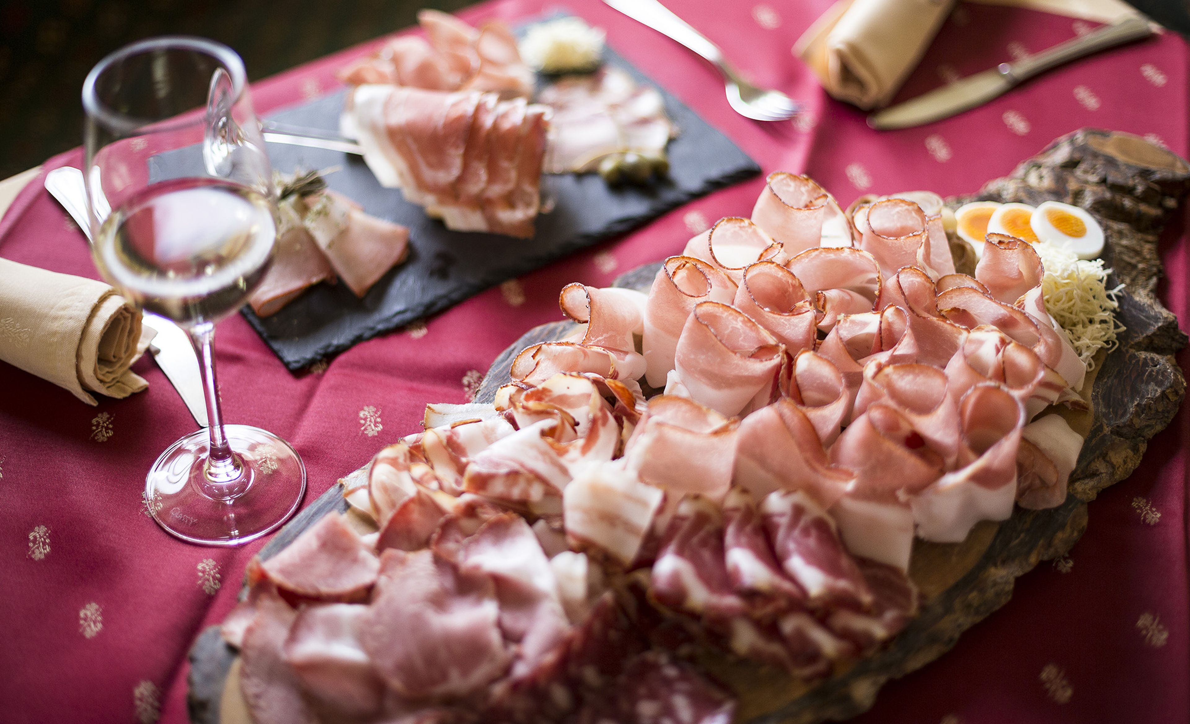 Eine Speck-Platte auf einem Holzbrett, dazu ein Glas Weißwein auf einer roten Tischdecke.