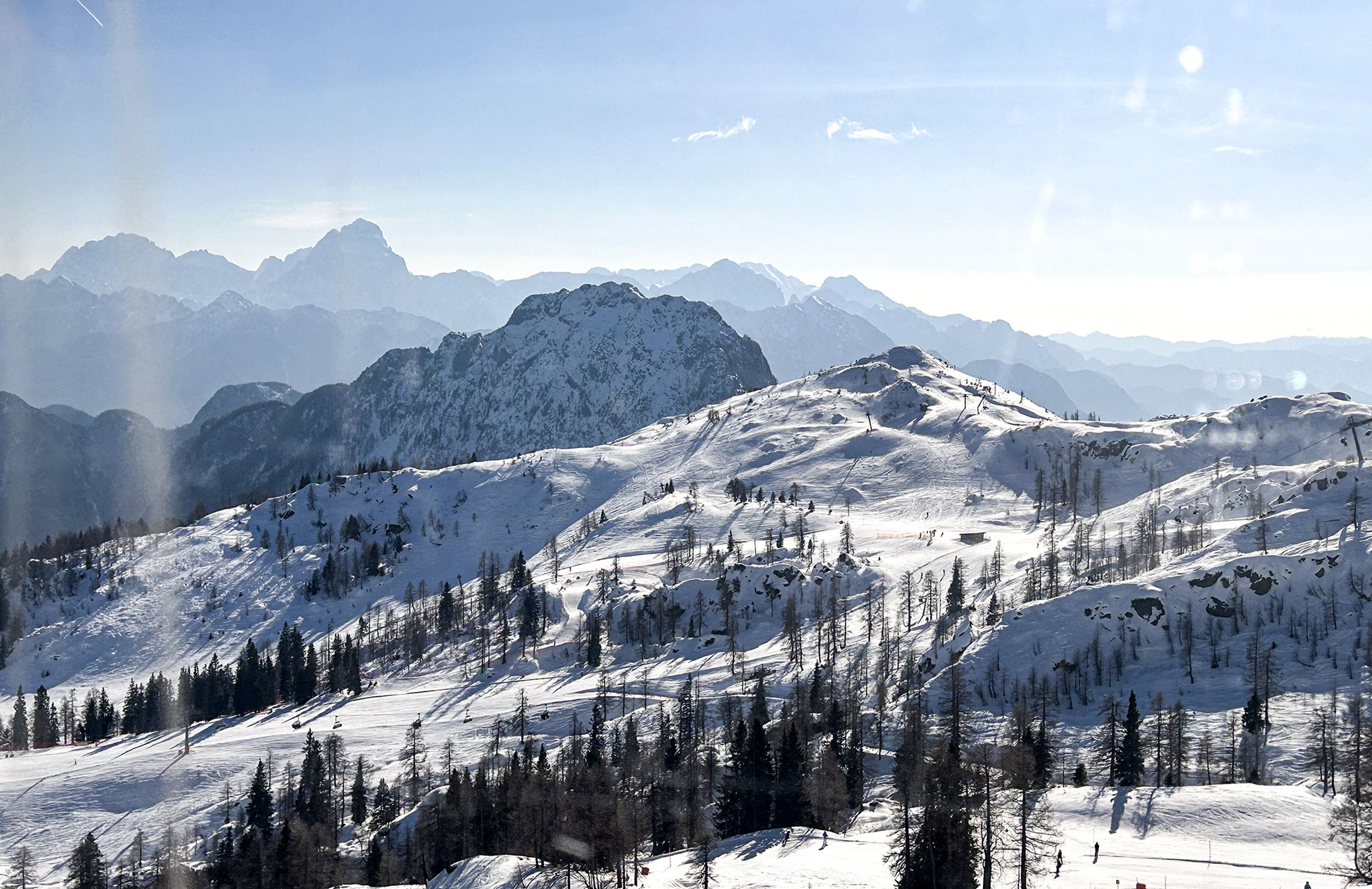 Schneebedeckte Berglandschaft mit vereinzelten Bäumen und fernen Gipfeln unter einem klaren blauen Himmel.
