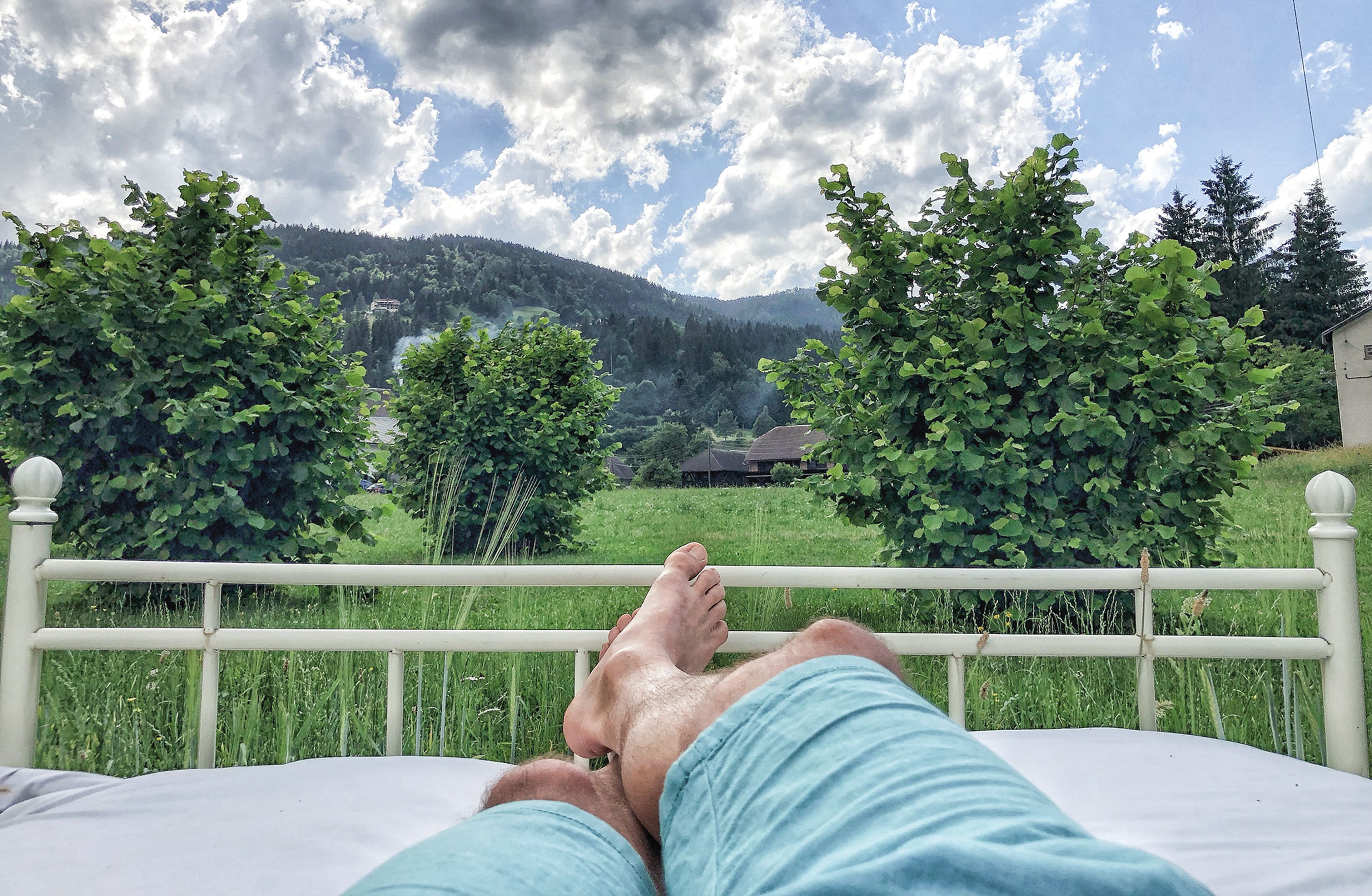 Person, die im Freien auf einem Bett liegt, blaue Shorts trägt und im Hintergrund einen Blick auf grüne Bäume, Berge und einen bewölkten Himmel hat.