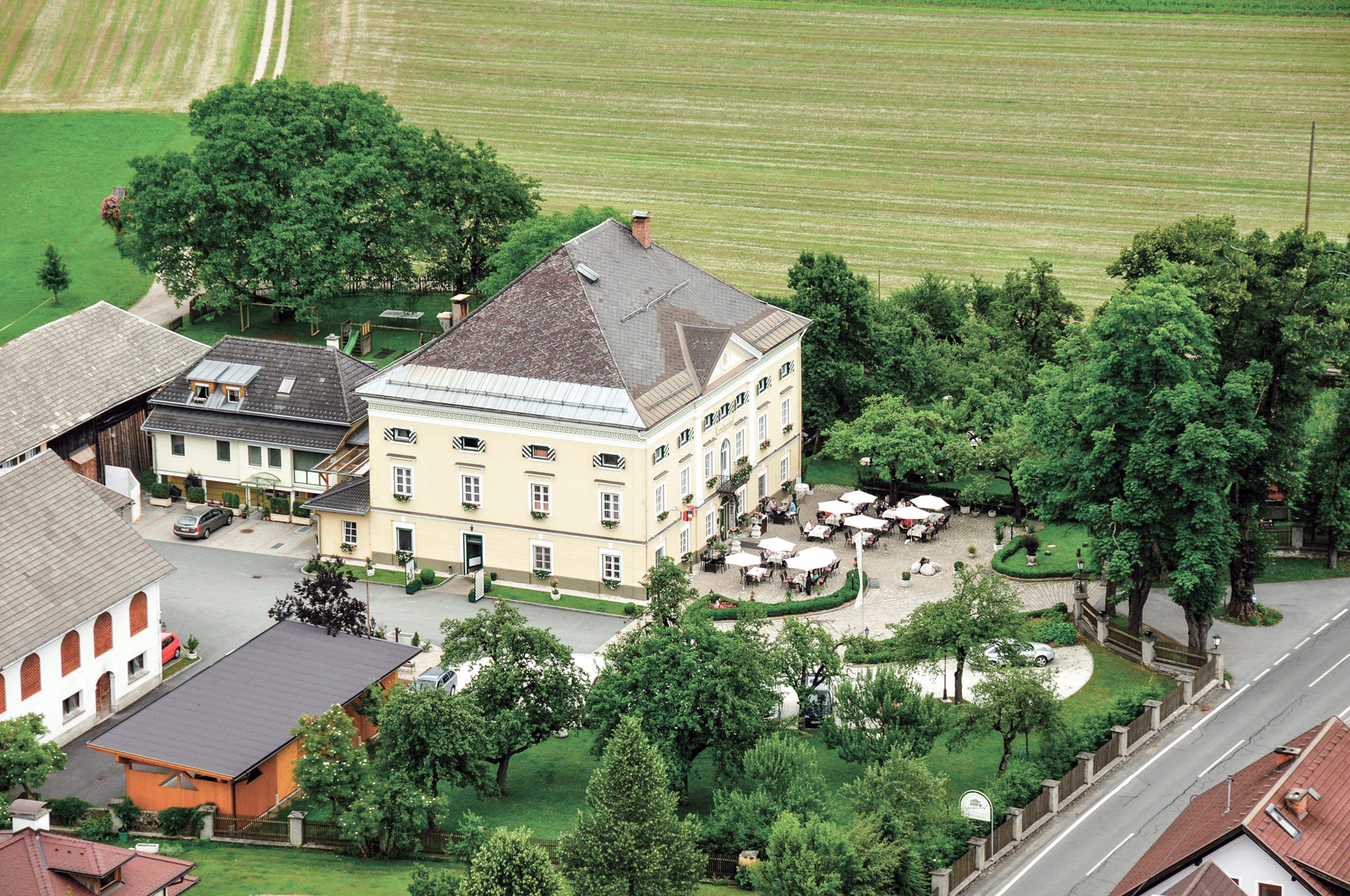 Luftaufnahme des großen, historischen Gebäudes Schloss Lerchenhof mit Innenhof, umgeben von Bäumen und Feldern, mit Sitzgelegenheiten im Freien und nahe gelegenen Wiese, Feldern und Bäumen