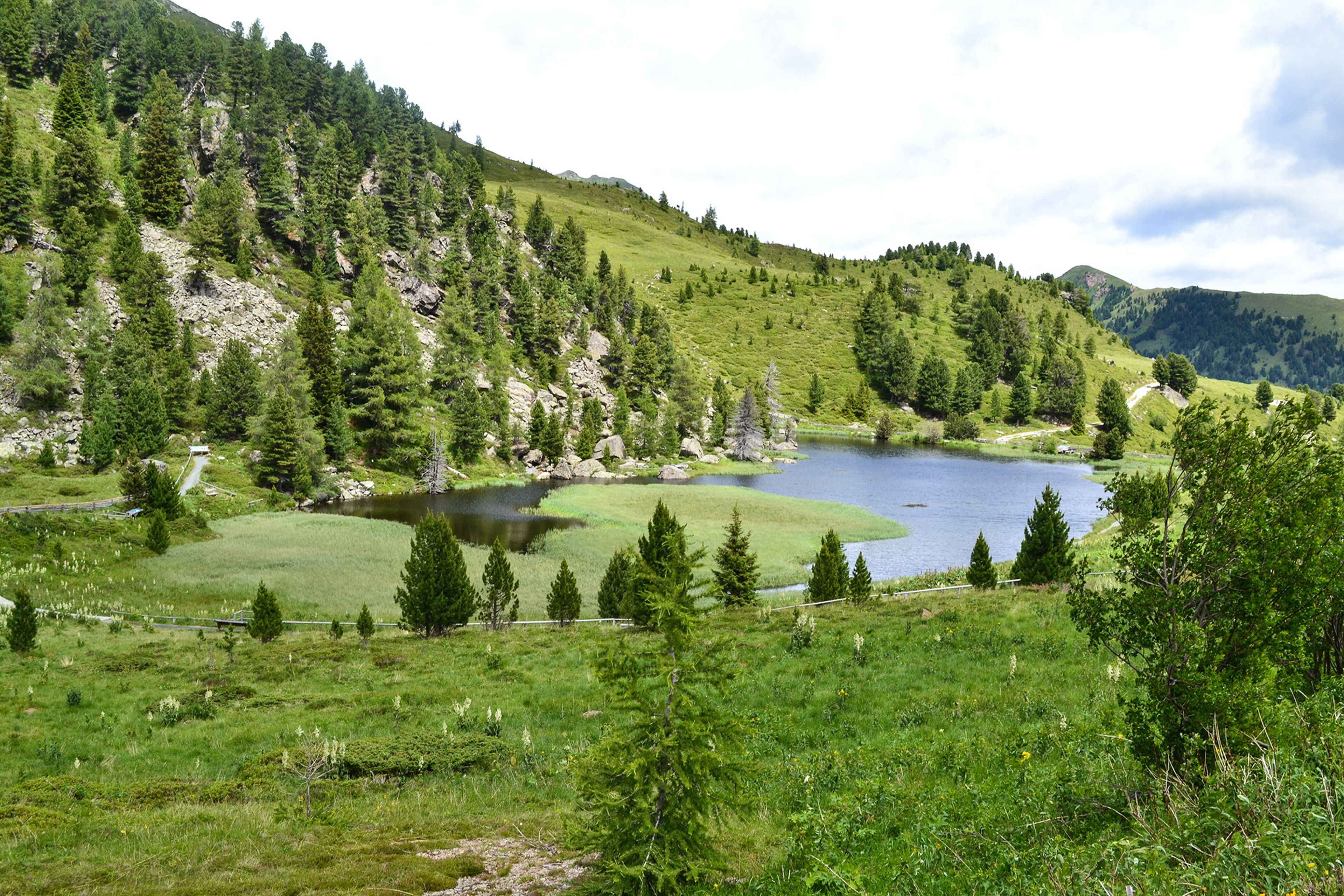 Eine ruhige Berglandschaft mit einem kleinen See, umgeben von üppigem grünem Gras, Bäumen und felsigen Hügeln unter einem teilweise bewölkten Himmel.