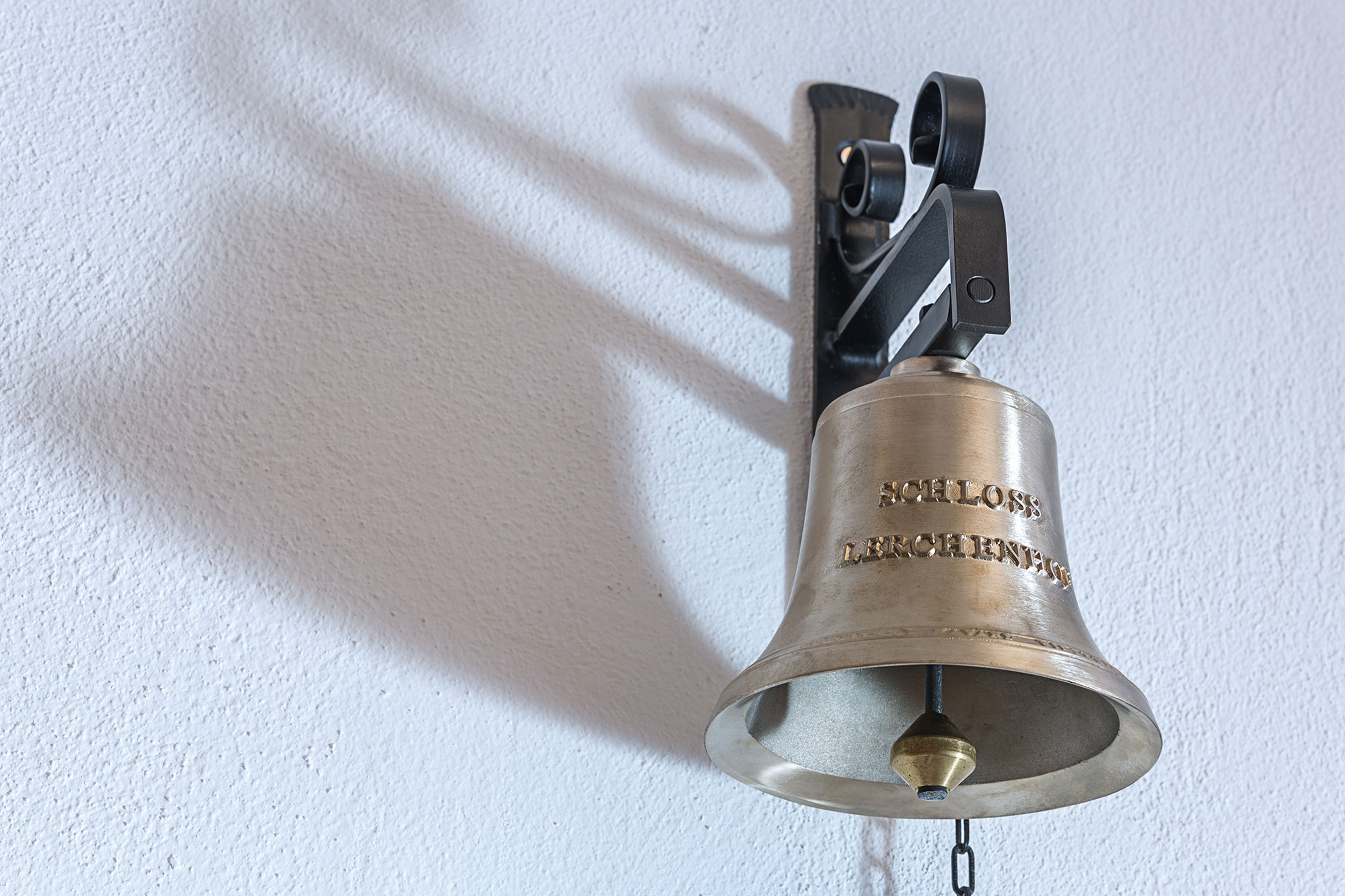 A brass bell mounted on a wall with a decorative bracket, casting a shadow. The bell has engraved text and a chain hanging below.
