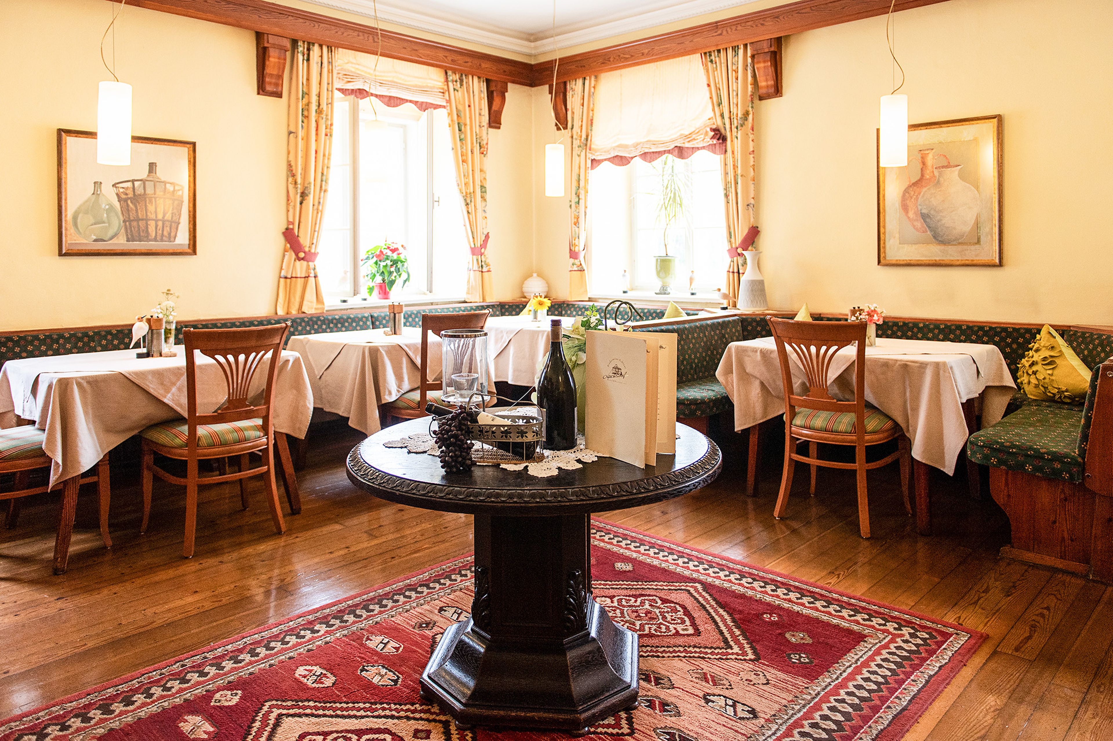 Ein gemütliches Restaurant-Interieur mit Holzböden, Tischen mit weißen Tischdecken, einem zentralen runden Tisch mit Dekor und Gemälden an den Wänden.