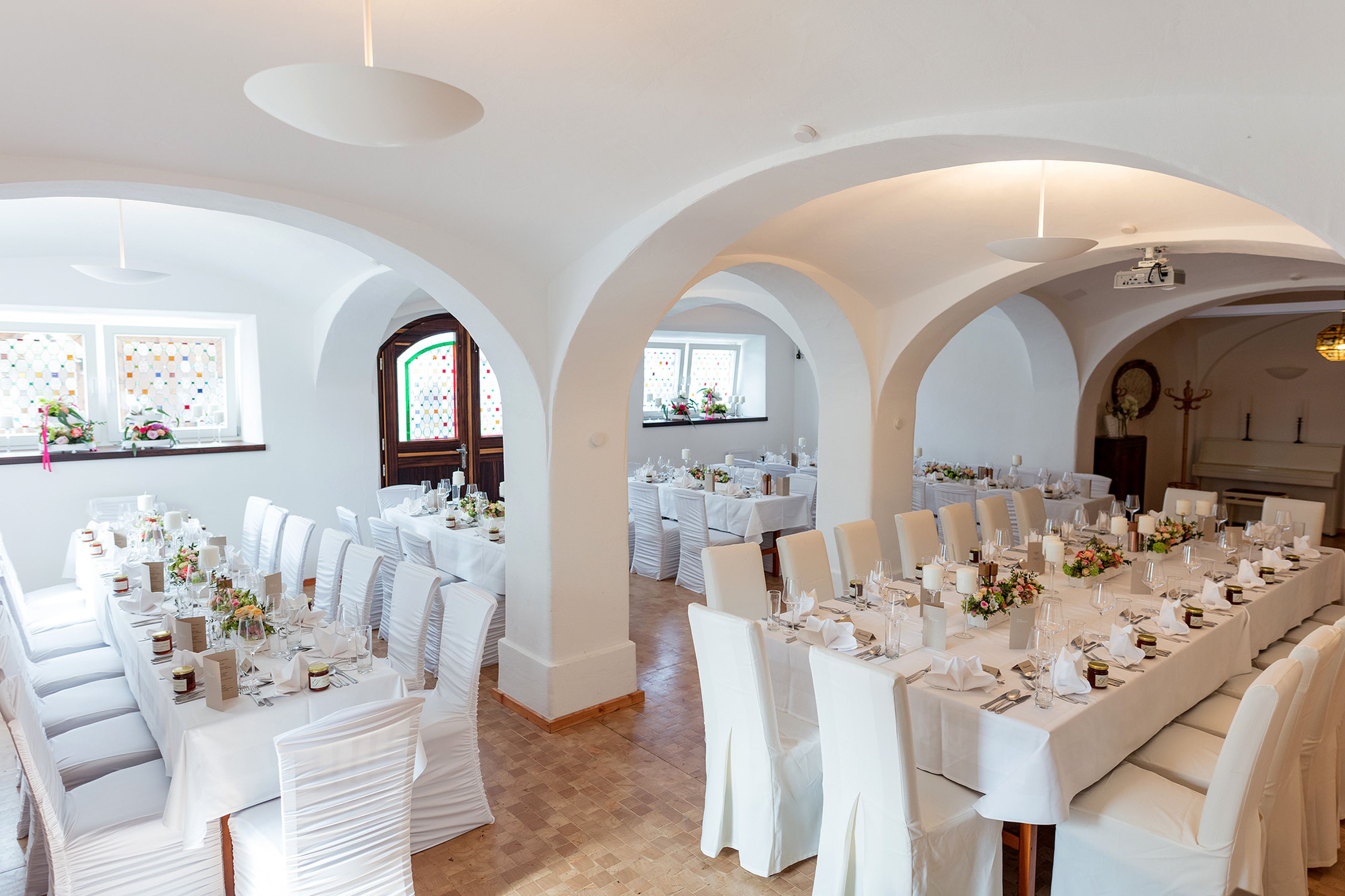 Eleganter Bankettsaal mit weißen Tischdecken und Stühlen, gedeckt für eine Veranstaltung. Torbögen und florale Tischdekorationen schaffen eine gehobene Atmosphäre.