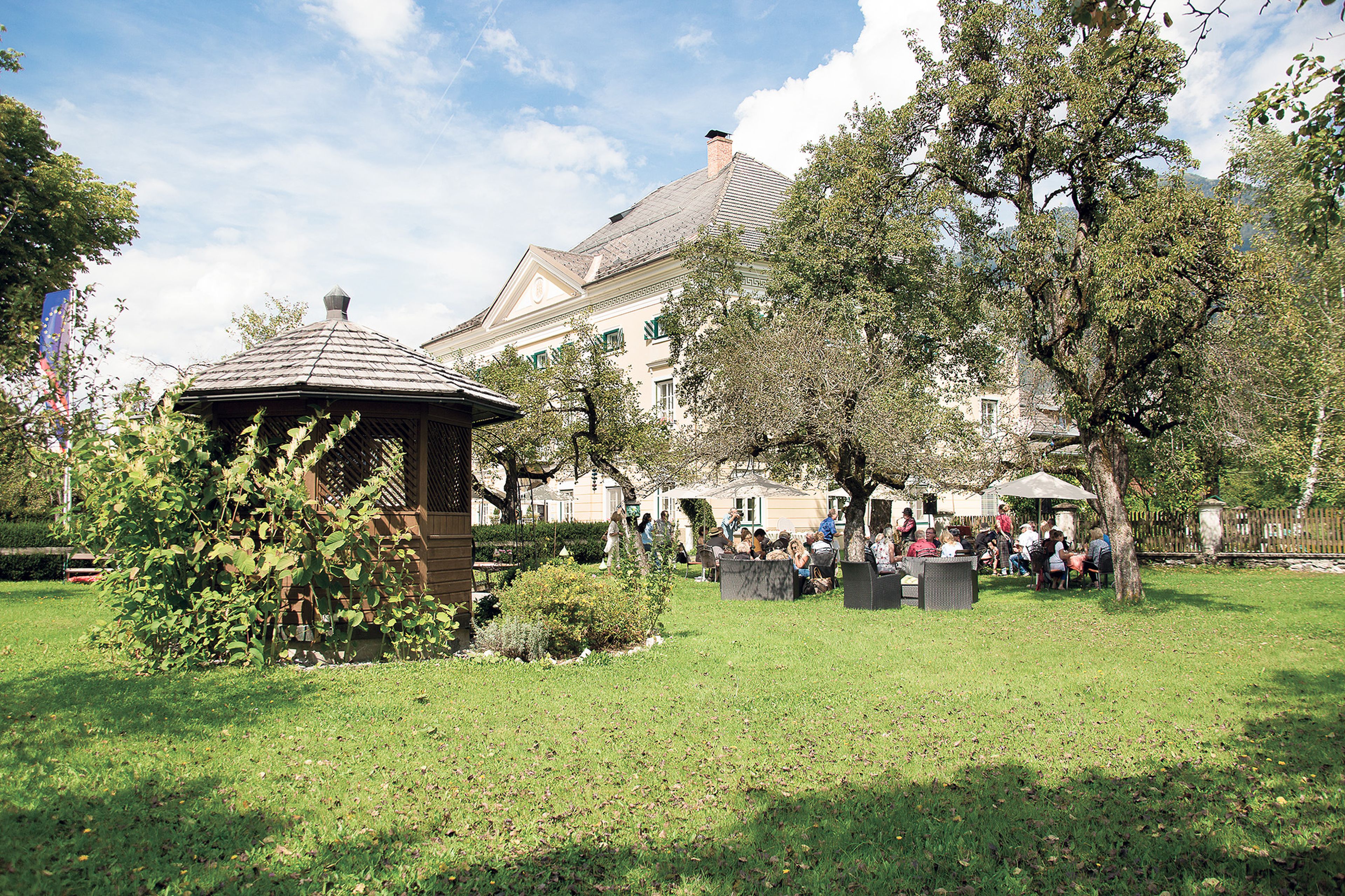 Eine Gruppe von Menschen sitzt auf Gartenmöbeln im Garten des Schloss Lerchenhof mit Bäumen und einem hölzernen, gemütlichen Pavillon